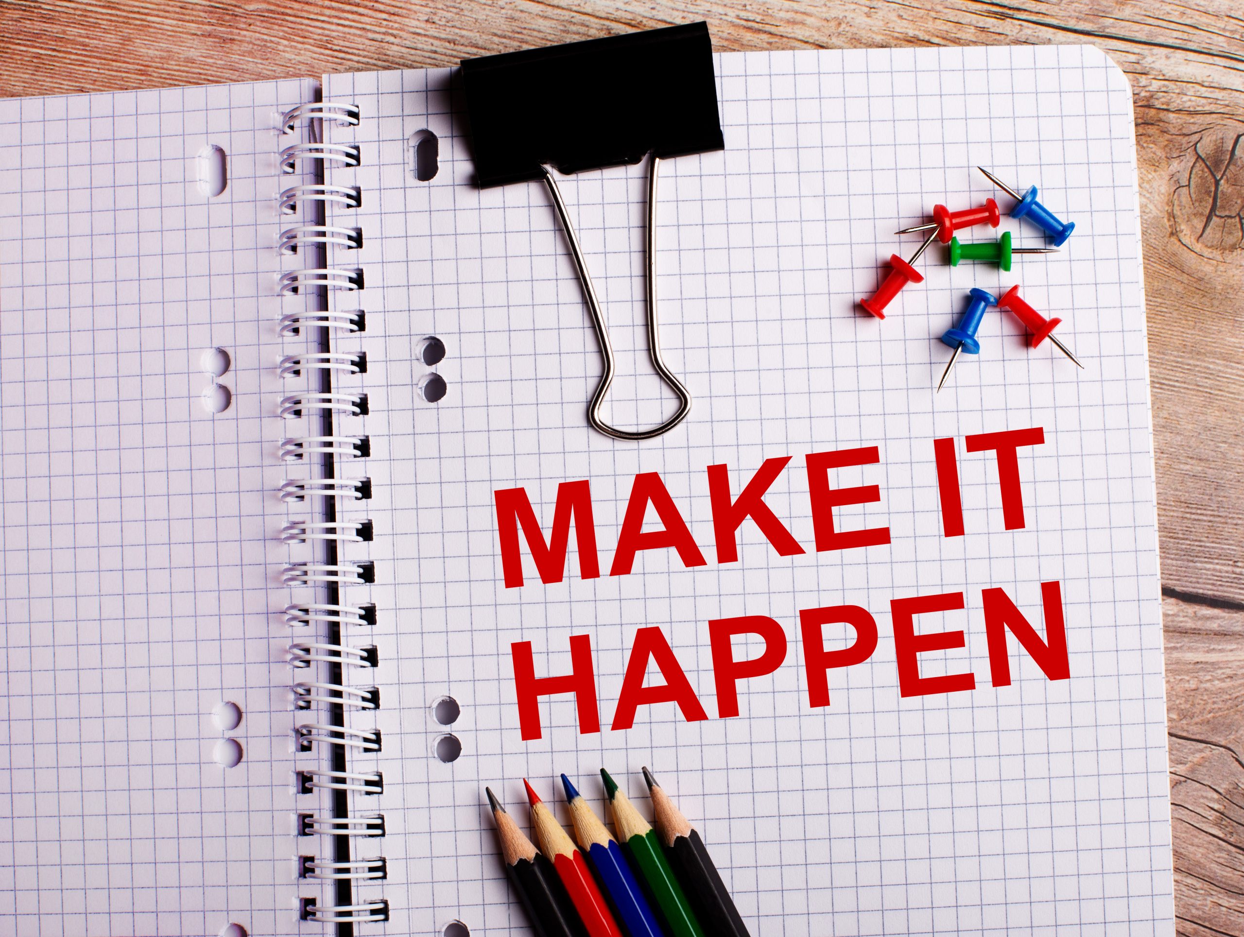 The words MAKE IT HAPPEN is written in a notebook near multi-colored pencils and buttons on a wooden background.